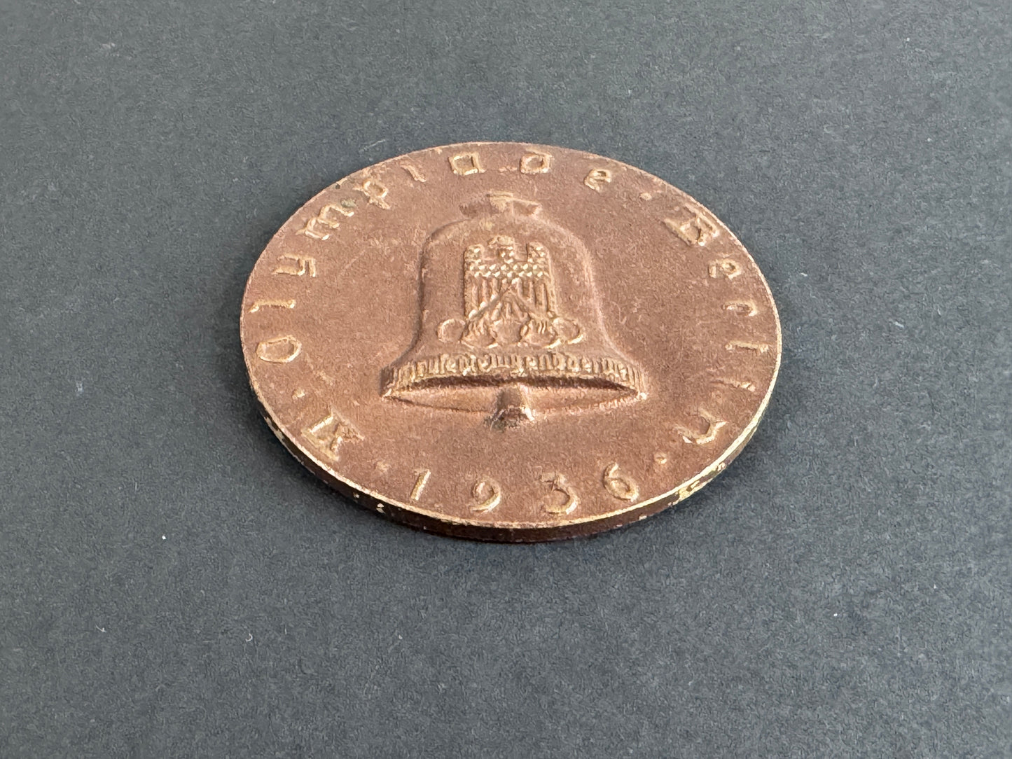 Bronze coin with a bell design on a gray background. For 9th Olympiad Berlin 1936.
