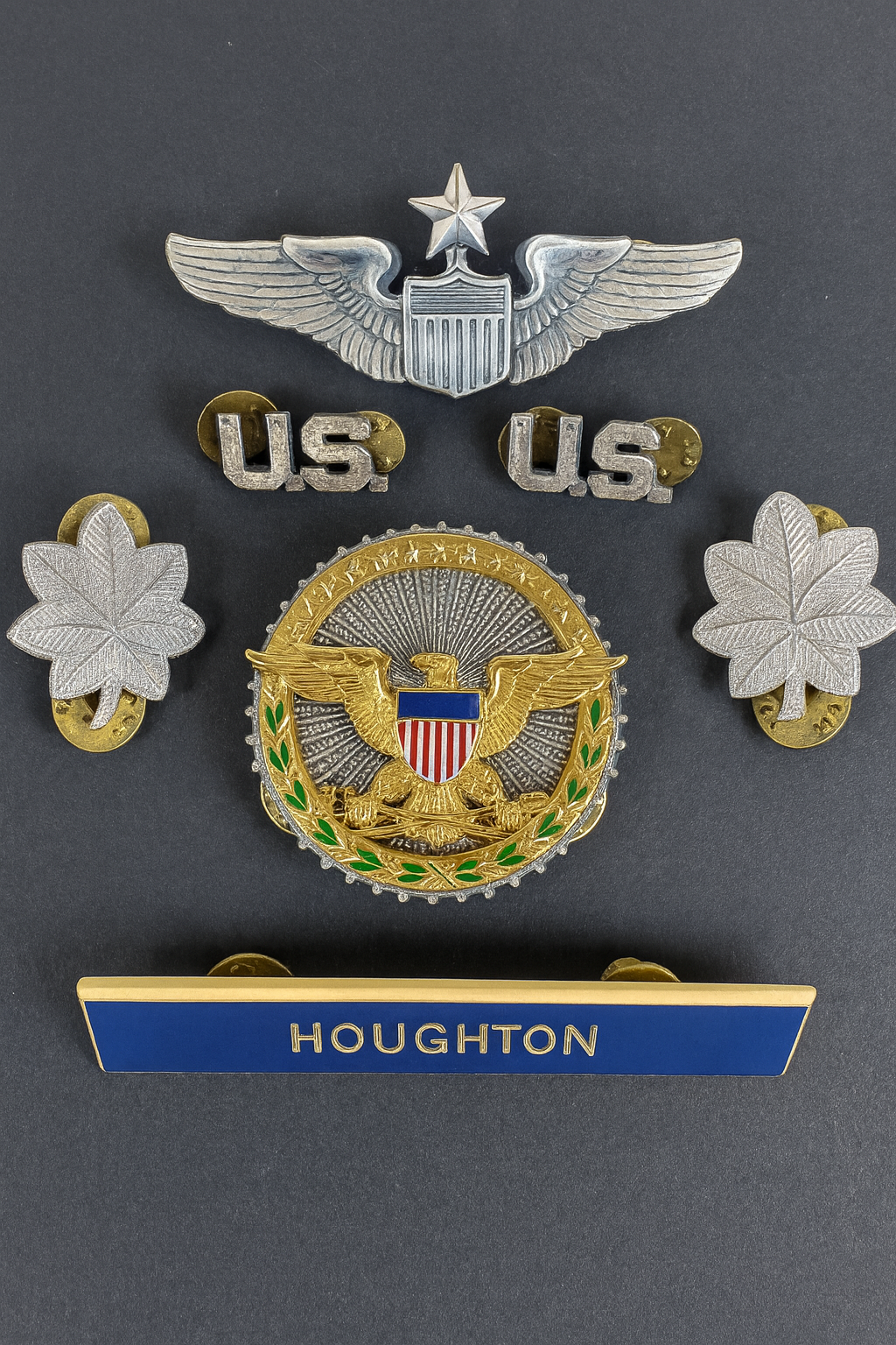 U.S. military insignia including a colonel’s rank leaves, Air Force command pilot wings, Great Seal cap badge, “U.S.” pins, and HOUGHTON nameplate on dark background.