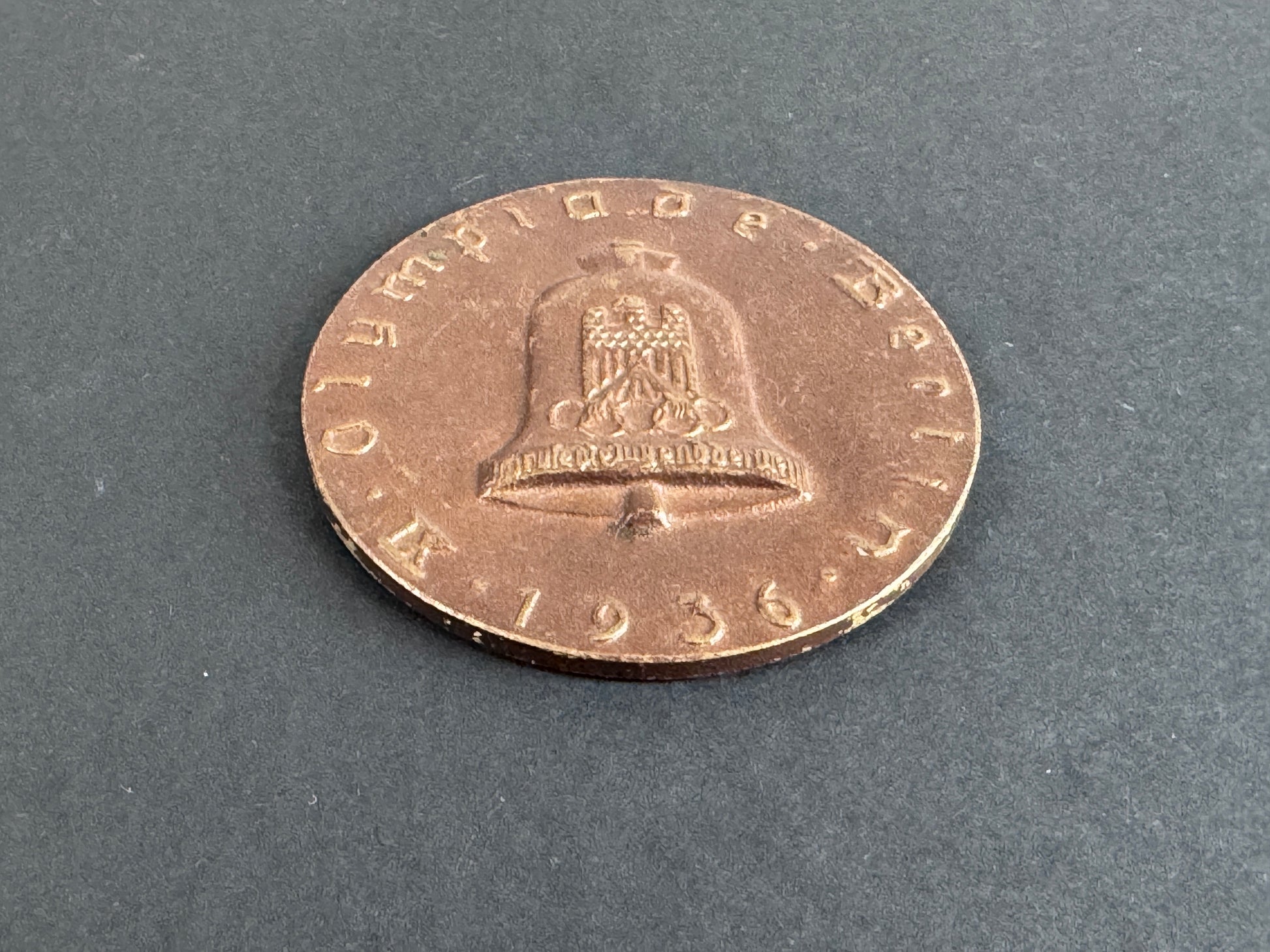 Bronze coin with a bell design on a gray background. For 9th Olympiad Berlin 1936.