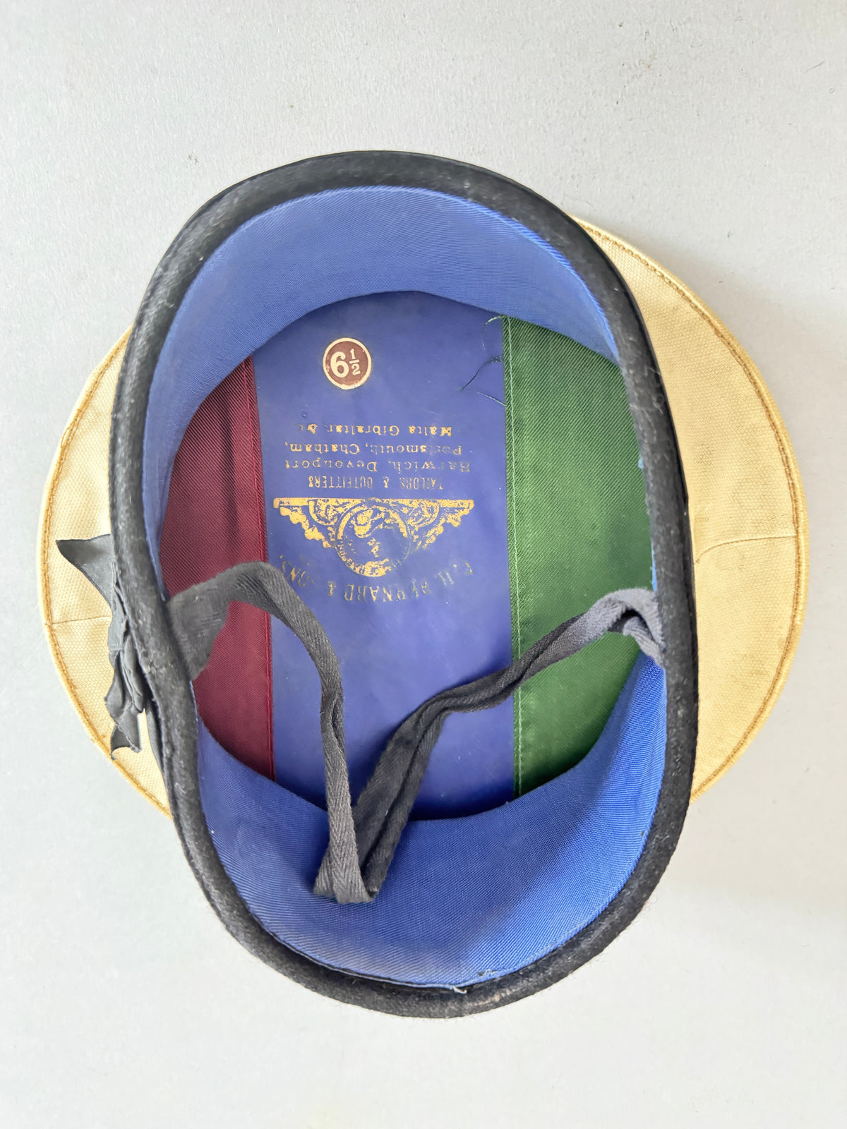 An overhead view of a gold-colored military helmet with a blue and green fabric lining. The interior features a blue panel with gold printed insignia and text, along with a red stripe at the top, and a gray chin strap.